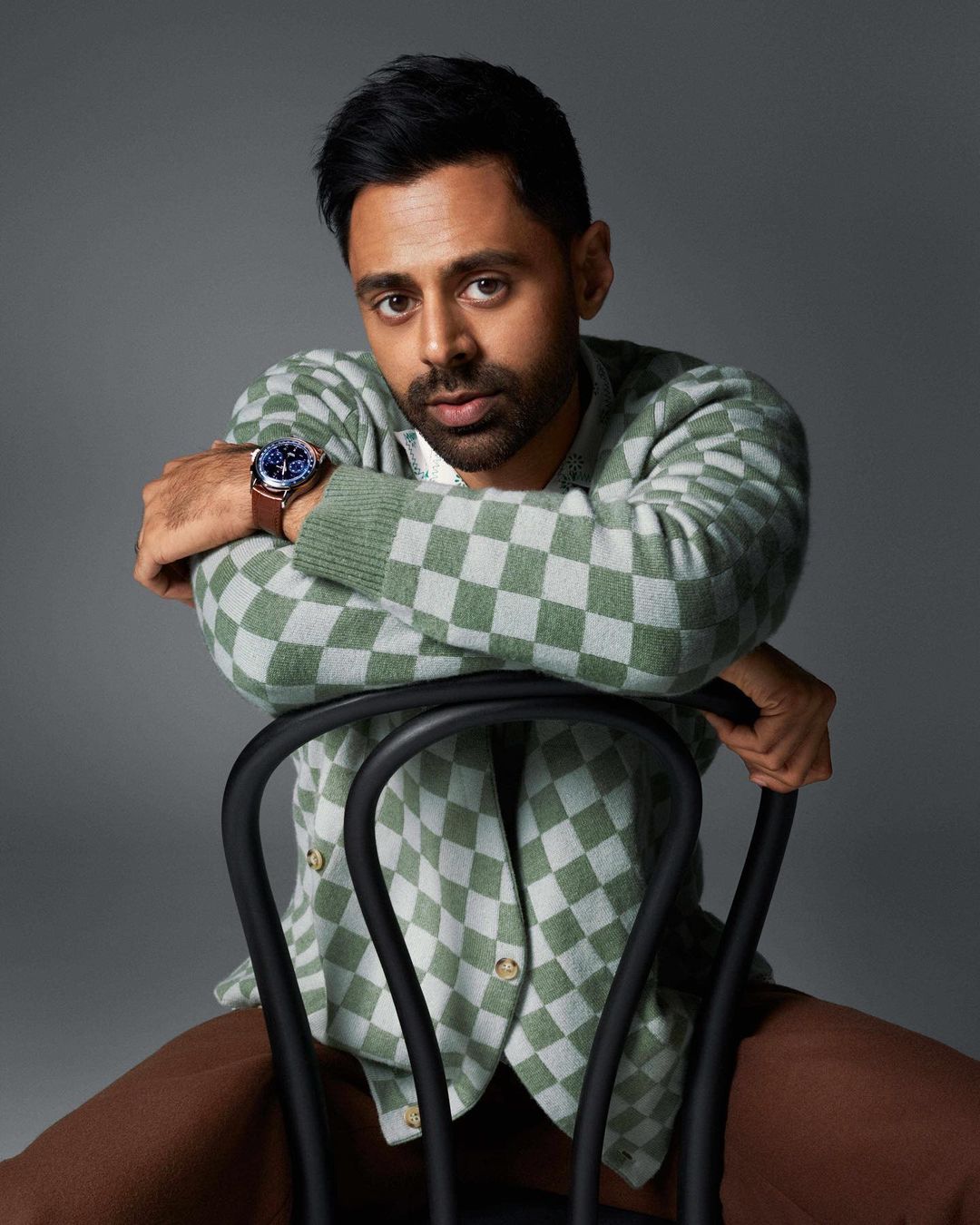 Portrait of a man in a checkerboard cardigan sitting backwards on a chair against a gray background in a studio setting.