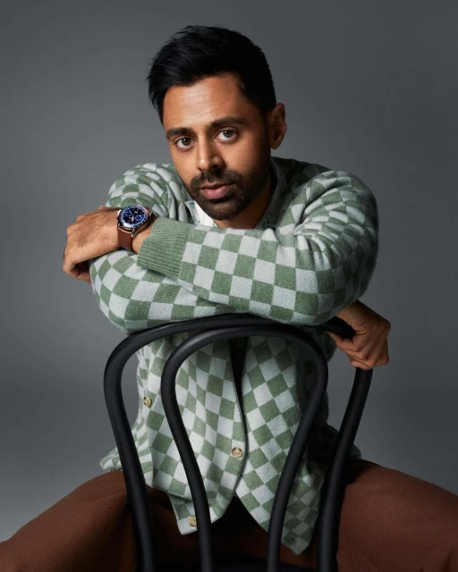 Portrait of a man in a checkerboard cardigan sitting backwards on a chair against a gray background in a studio setting.