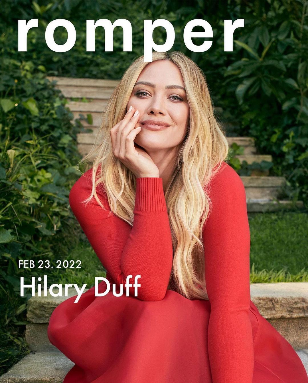 Blonde woman, Hilary Duff, smiles on Romper cover in a red dress, with a green foliage background and natural lighting.