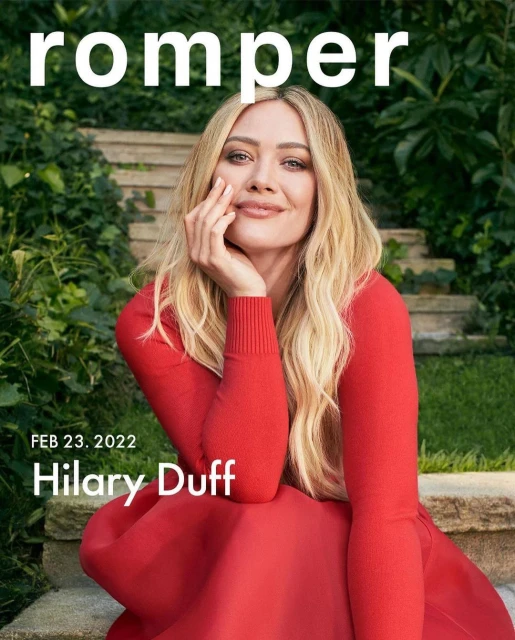 Blonde woman, Hilary Duff, smiles on Romper cover in a red dress, with a green foliage background and natural lighting.