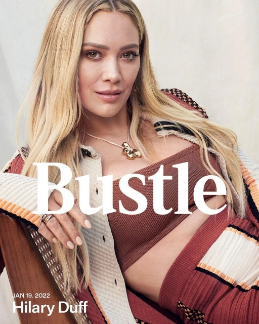 Blonde woman, Hilary Duff, in a portrait wearing a brown top and sweater, with the word Bustle in large white font overlayed.