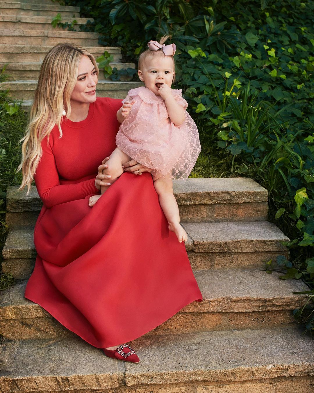 A woman in a red dress sits on stone steps holding a baby girl in a pink dress, with a lush green leafy background.