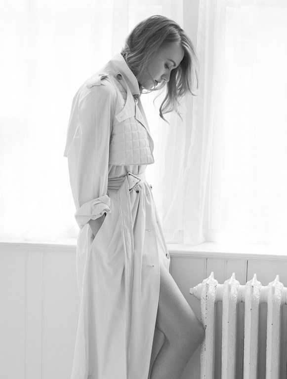 Monochrome portrait of a stylish adult woman in a trench coat posing indoors by a window with flowing curtains and a vintage radiator.