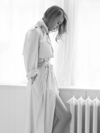 Monochrome portrait of a stylish adult woman in a trench coat posing indoors by a window with flowing curtains and a vintage radiator.