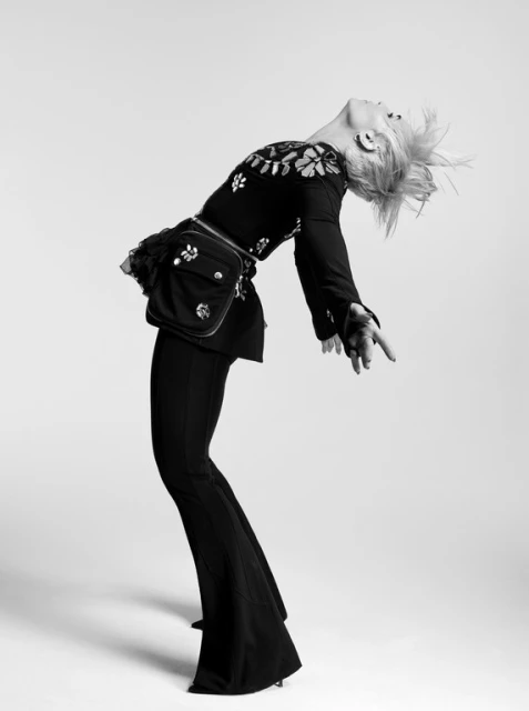 Black-and-white portrait of a woman in flared pants, a floral-patterned top, and a bum bag, leaning backward in a photo studio.