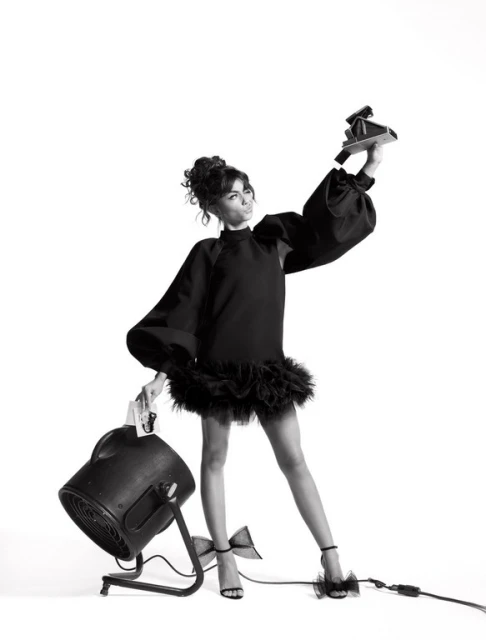 Elegant woman in black dress holding vintage camera and light in a studio fashion shot with a clean white backdrop, glamorous and stylish.