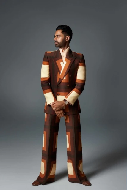 Full-length portrait of a man in a patterned suit looking to the left against a grey background in a fashion editorial style.