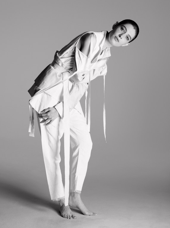 Woman poses in white linen suit with tie detail on a gray background in a studio shot. Fashion photography in black and white.
