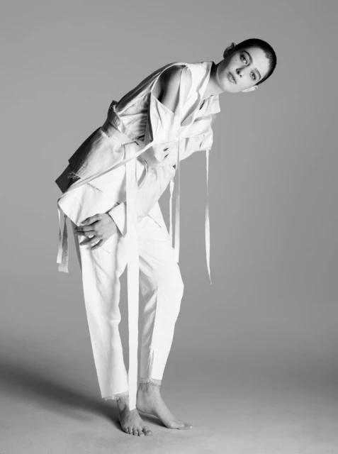 Woman poses in white linen suit with tie detail on a gray background in a studio shot. Fashion photography in black and white.