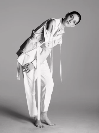 Woman poses in white linen suit with tie detail on a gray background in a studio shot. Fashion photography in black and white.