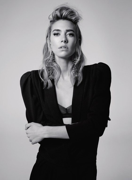 Portrait of a woman with braided updo wearing structured black blazer and ornate crystal earrings against light grey background.