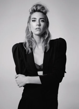 Portrait of a woman with braided updo wearing structured black blazer and ornate crystal earrings against light grey background.