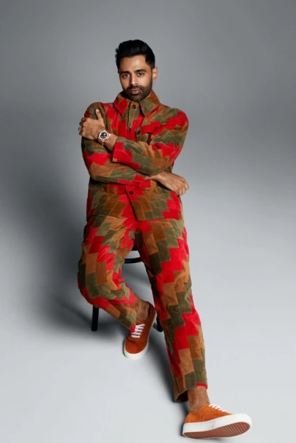 Adult male sitting indoors, wearing a patterned multicolor suit and brown sneakers, striking a confident pose in a studio setting.