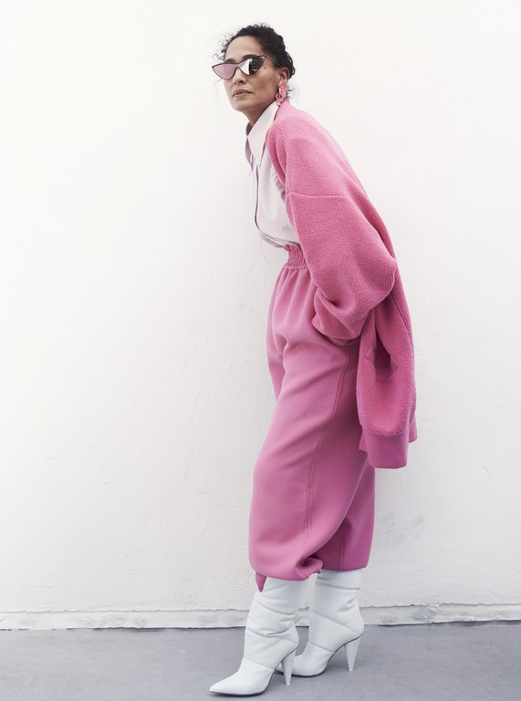 Woman in oversized pink outfit poses against white wall. She wears sunglasses and white heeled boots in a fashion style shot.