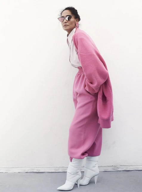Woman in oversized pink outfit poses against white wall. She wears sunglasses and white heeled boots in a fashion style shot.