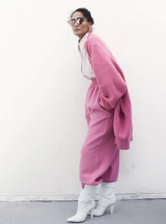 Woman in oversized pink outfit poses against white wall. She wears sunglasses and white heeled boots in a fashion style shot.