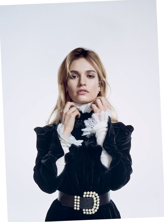 Blonde woman in a black velvet dress with a white ruffled collar and pearl belt against a white background in a studio shot.