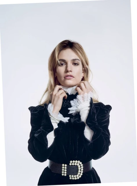 Blonde woman in a black velvet dress with a white ruffled collar and pearl belt against a white background in a studio shot.
