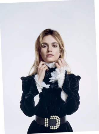 Blonde woman in a black velvet dress with a white ruffled collar and pearl belt against a white background in a studio shot.