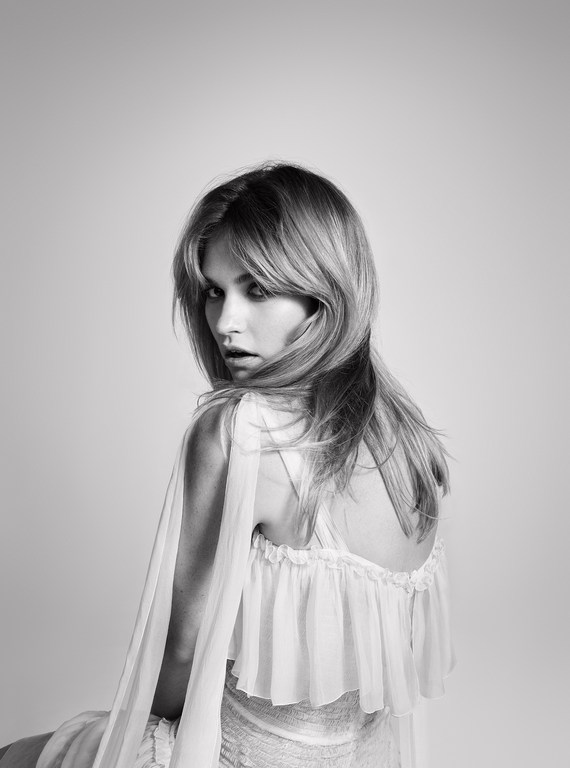Black and white portrait of an adult female with blonde hair, wearing a ruffled white dress, looking over her shoulder with an open mouth.