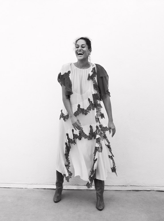 Black and white portrait of a smiling woman in a patterned dress and boots, standing against a neutral background in a photo studio.