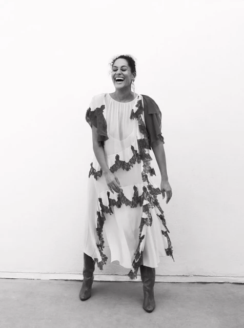 Black and white portrait of a smiling woman in a patterned dress and boots, standing against a neutral background in a photo studio.