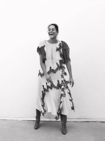 Black and white portrait of a smiling woman in a patterned dress and boots, standing against a neutral background in a photo studio.