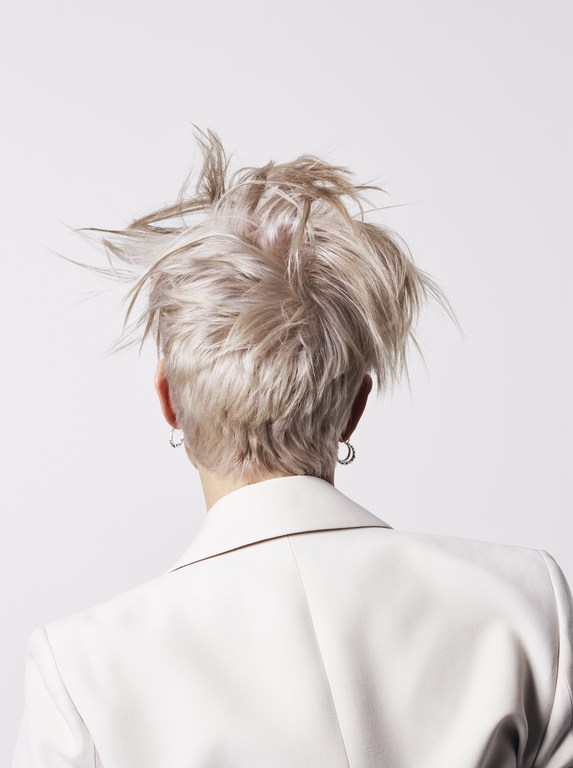 Rear view of a person with tousled blonde hair wearing a white blazer against a white background.