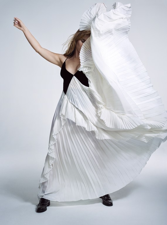 Fashionable woman in a black and white dress with pleated details is in a light studio setting, posing dynamically with arm raised.