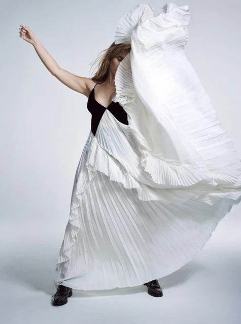 Fashionable woman in a black and white dress with pleated details is in a light studio setting, posing dynamically with arm raised.