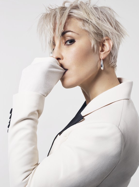 Blonde adult female in a white suit and gloves poses in a studio with eye-level angle and shallow depth of field. Portrait orientation.