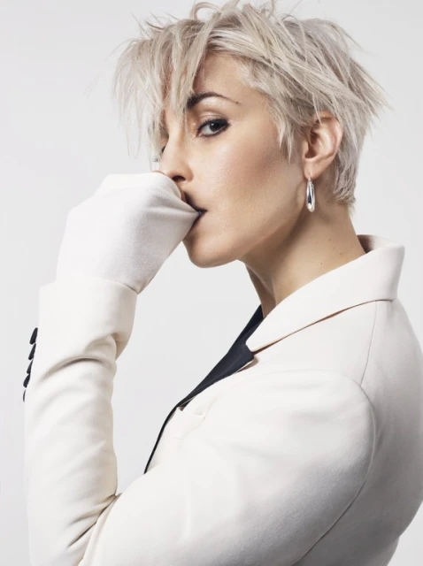 Blonde adult female in a white suit and gloves poses in a studio with eye-level angle and shallow depth of field. Portrait orientation.