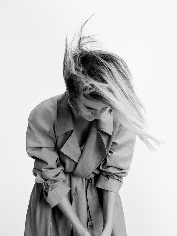 Monochrome portrait of a woman wearing a belted trench coat with her hair tossed dynamically against a plain white background.