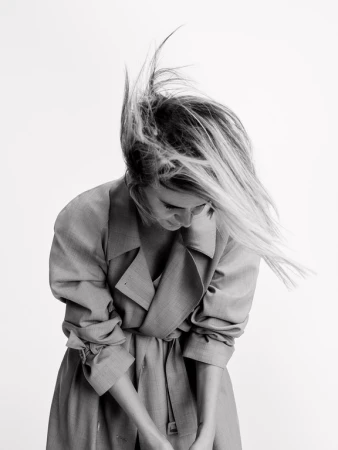 Monochrome portrait of a woman wearing a belted trench coat with her hair tossed dynamically against a plain white background.