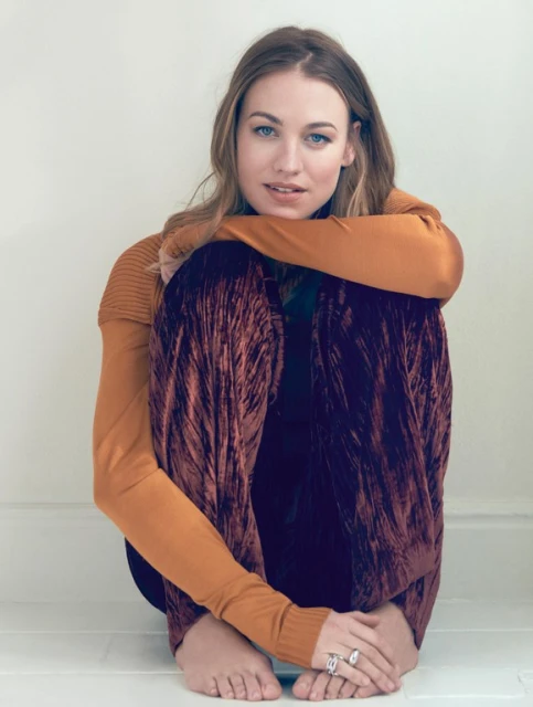 Young woman with long wavy hair, wearing a rust-colored top and patterned velvet pants, sitting with her arms around her legs.