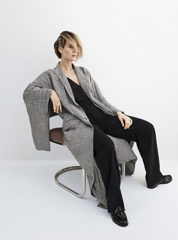 Woman with short blonde hair seated on a chair wearing a grey tweed coat and black trousers by Kerry Hallihan