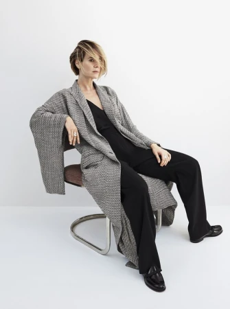 Woman with short blonde hair seated on a chair wearing a grey tweed coat and black trousers by Kerry Hallihan