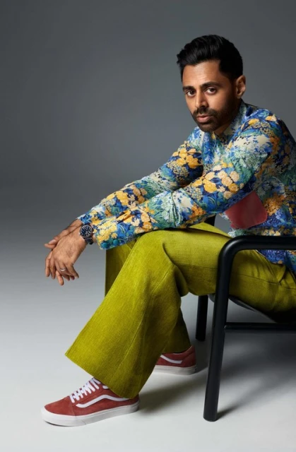 Adult male in floral shirt and green pants sits on chair in studio, wearing red sneakers and a watch, looking at the camera.