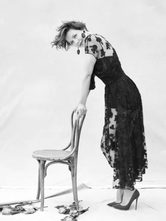 Monochrome portrait of a woman leaning on a wooden chair wearing a dark floral lace dress against a plain background