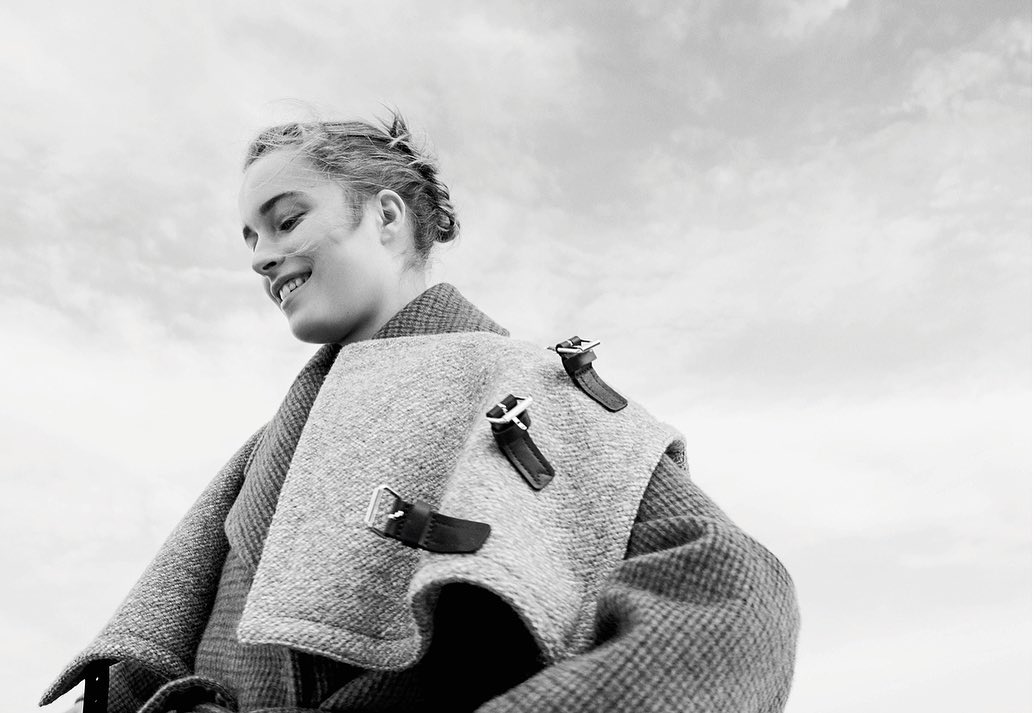 Monochrome portrait of a smiling young woman with braided hair wearing a grey wool coat with leather buckle details against cloudy sky.