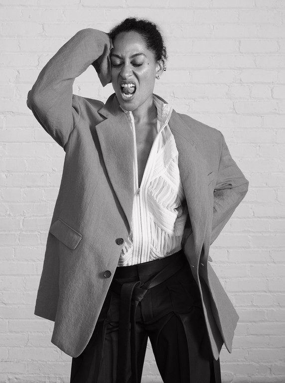 A woman in a blazer and zip-up shirt poses against a brick wall, with one hand on her head and her mouth open in an expressive yell.