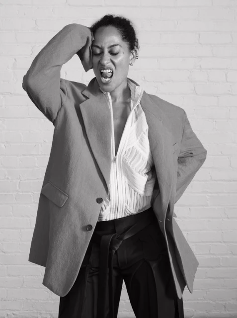 A woman in a blazer and zip-up shirt poses against a brick wall, with one hand on her head and her mouth open in an expressive yell.