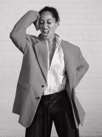A woman in a blazer and zip-up shirt poses against a brick wall, with one hand on her head and her mouth open in an expressive yell.