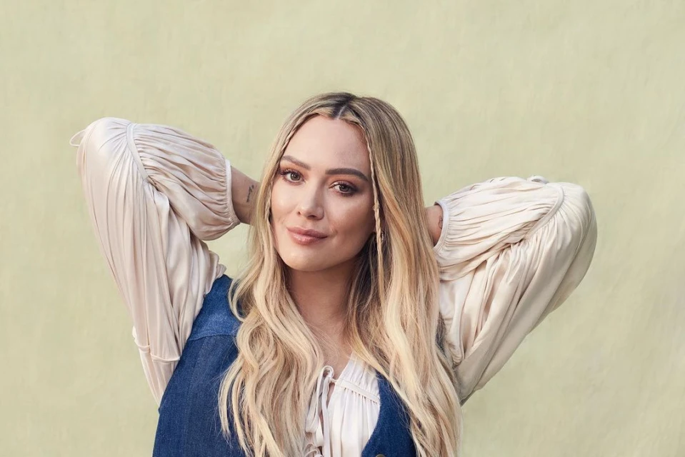 Close-up portrait of Hilary Duff with blonde wavy hair and denim vest, looking at the camera with a soft smile, set against a neutral backdrop.