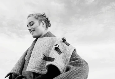 Monochrome portrait of a smiling woman wearing a textured wool coat outdoors against a cloudy sky.