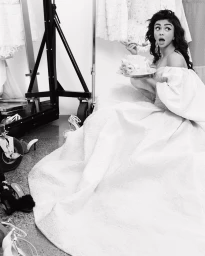 Black and white portrait of a woman in a large white off-the-shoulder gown holding a cake and a spoon with a surprised expression.