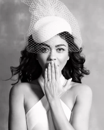 Black and white portrait of a woman wearing a white pillbox hat with birdcage veil and halter top, covering mouth with hand.