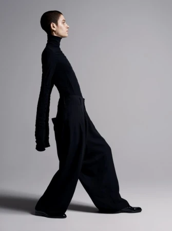 Model with a shaved head wearing a black turtleneck and wide trousers against a light grey seamless background