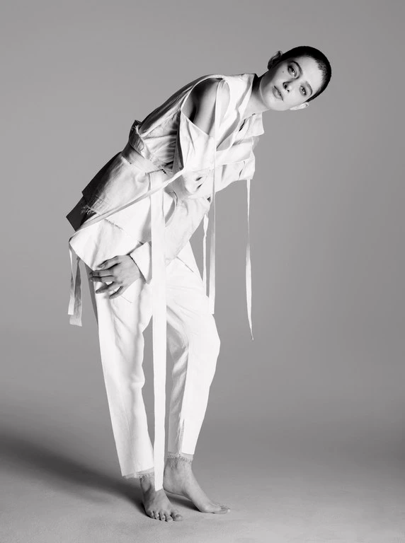 Model in white belted jacket and trousers leaning forward against a plain studio background.