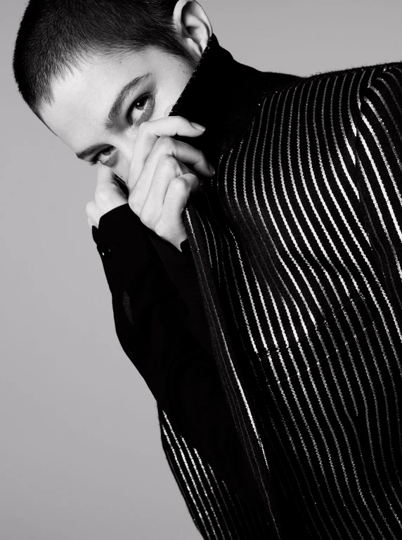 Close-up portrait of a model with short cropped hair pulling up a striped jacket collar to partially cover her face.
